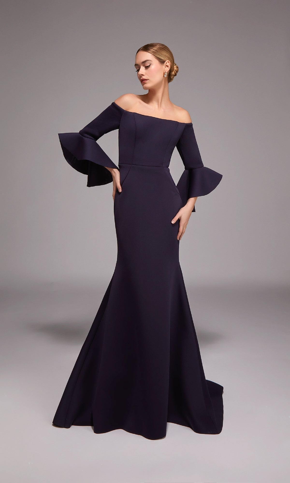 A woman poses in a studio wearing the Alyce 27777, a floor-length dark purple off-the-shoulder mermaid gown with flared bell sleeves.